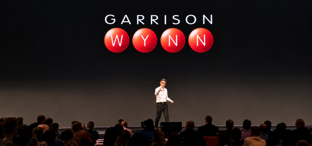 Garrison Wynn delivering a keynote on stage beneath his logo to a large audience